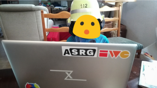 Little kid with a costume firemen helmet sitting infront of a laptop.