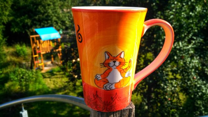 A mug in warm colours with a drawn cat meditating in a typical cross legged pose. A vibrant green garden is stretching out in the background. A play house for children peeks out behind some trees.